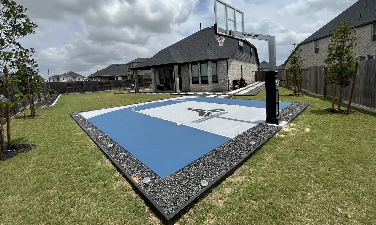 Blue and gray backyard basketball court with gravel border behind a home