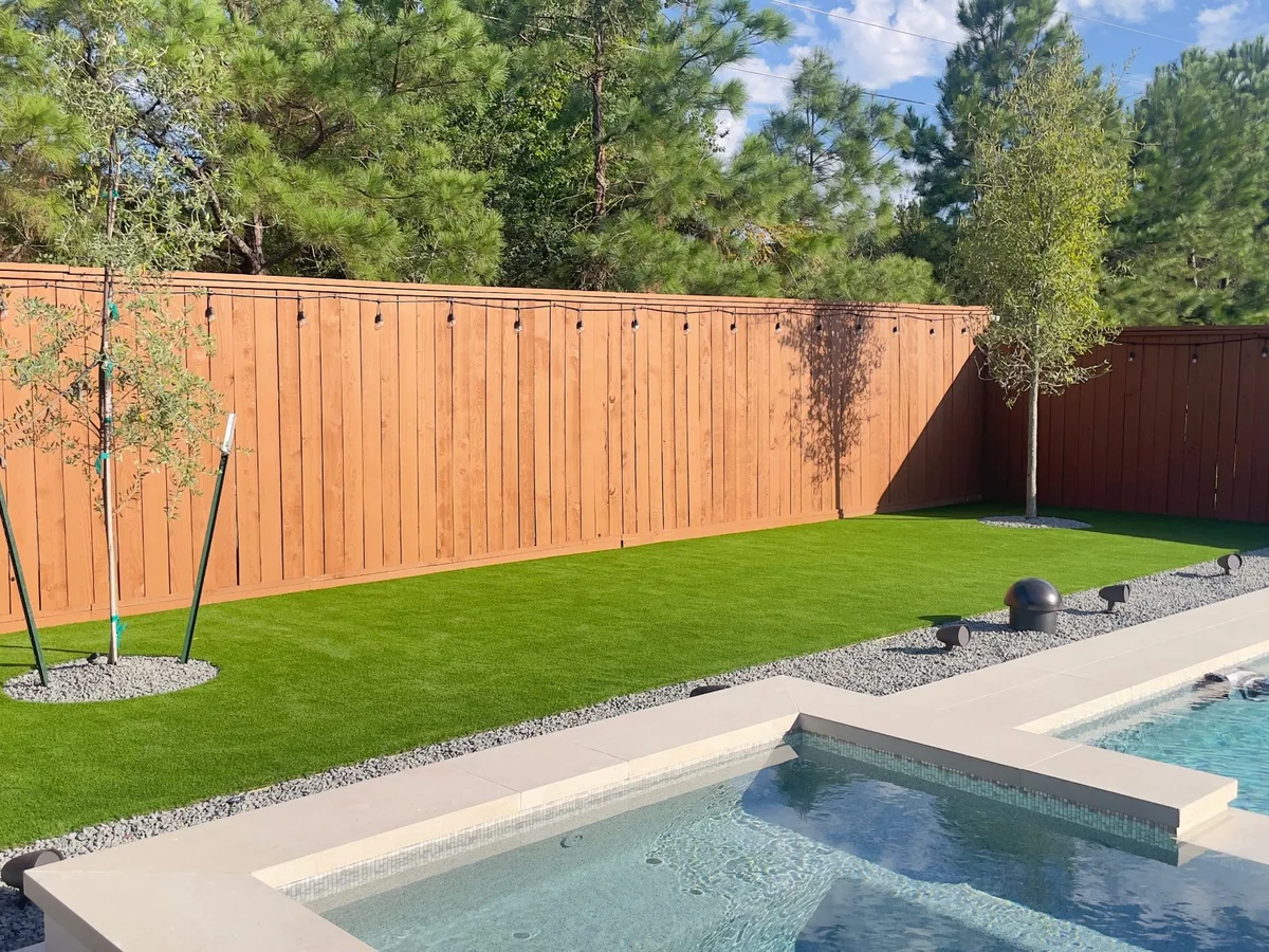 Artificial turf backyard with a wood fence, young tree and pool edge