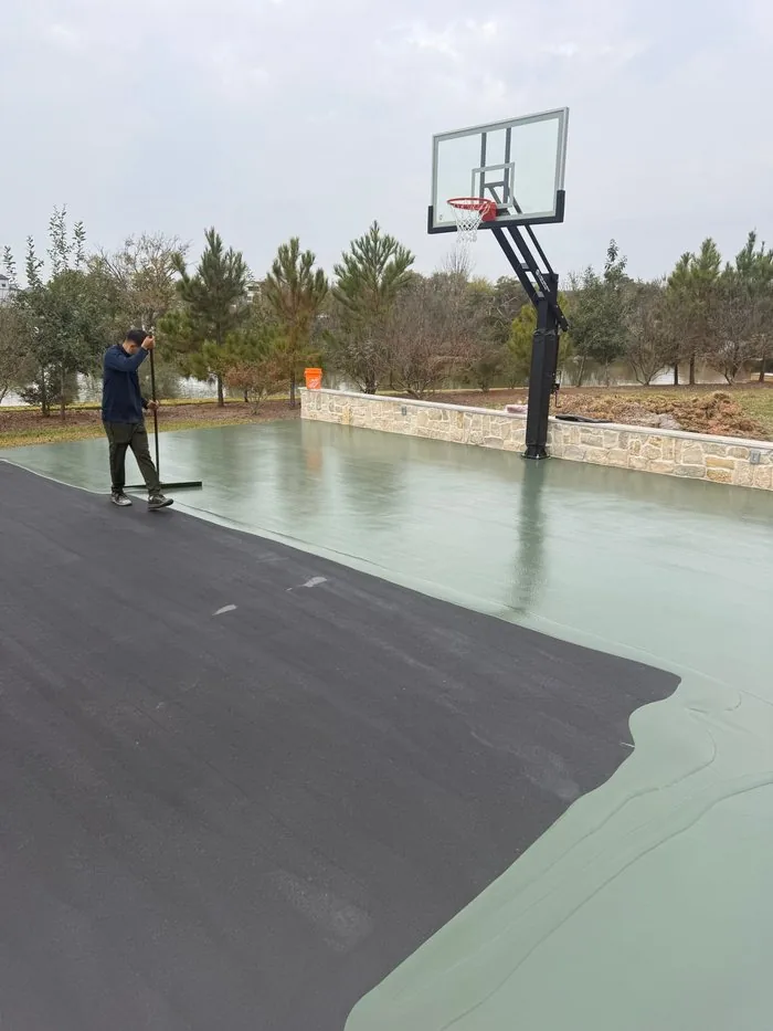 Crew member squeegeeing green coating over the dark resurfacer on a basketball court