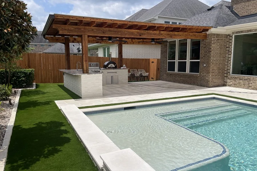 Wood pergola over an outdoor kitchen beside a pool with paver patio and turf